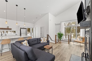 A modern interior with a grey sectional sofa, a kitchen with a stainless steel refrigerator, and a large window overlooking a forest. - Tremblant ski 8 min, Spa, Fireplace, Dog-Friendly (Lac-Supérieur)