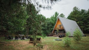A cabin with a sloped roof, surrounded by trees and a grassy area with outdoor seating. - Cozy A-frame w/HOT TUB, 5min walk to lake, King Bed, 30 min to Dells (Friendship)