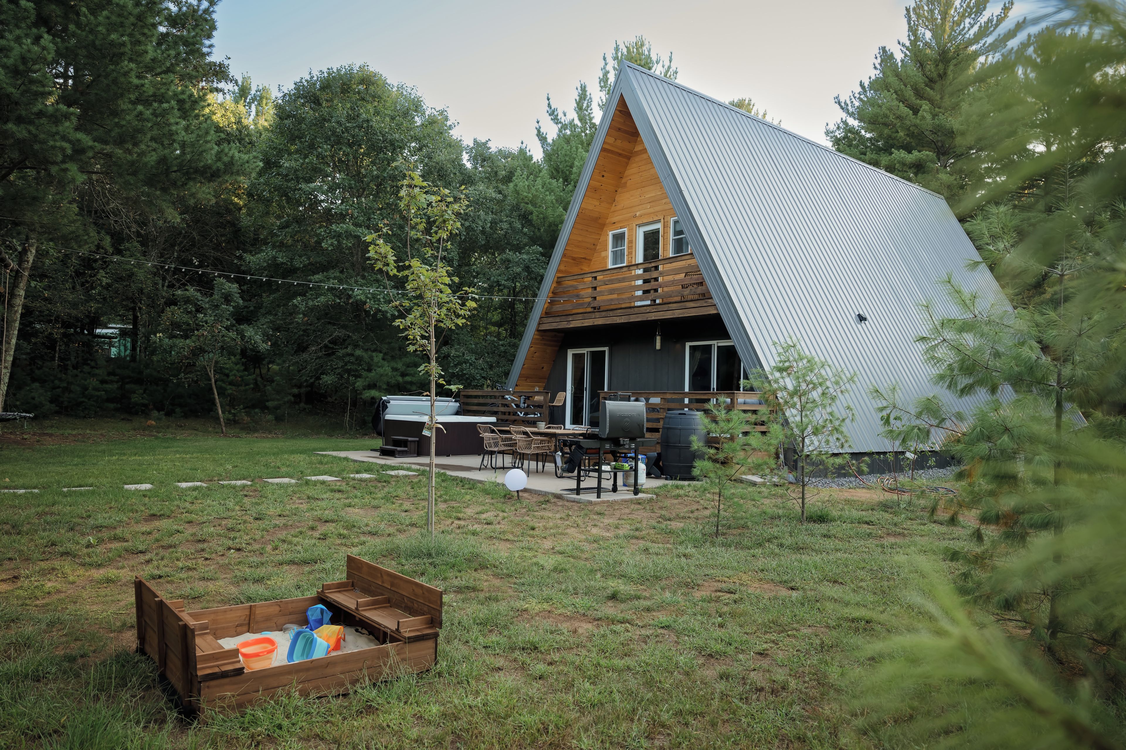 A modern A-frame house with a deck, outdoor furniture, and a sandbox.