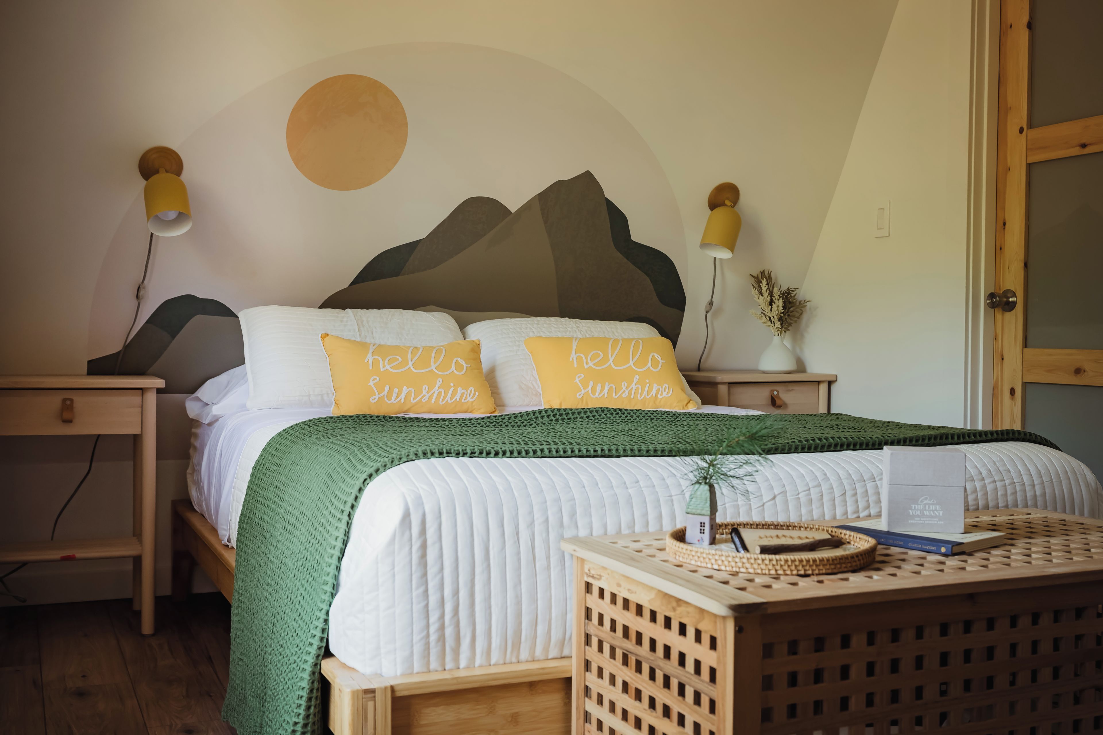 A neatly made bed with a green blanket, two pillows with 'hello Sunshine' text, a wall with a circular design, and a wooden nightstand with a lamp.