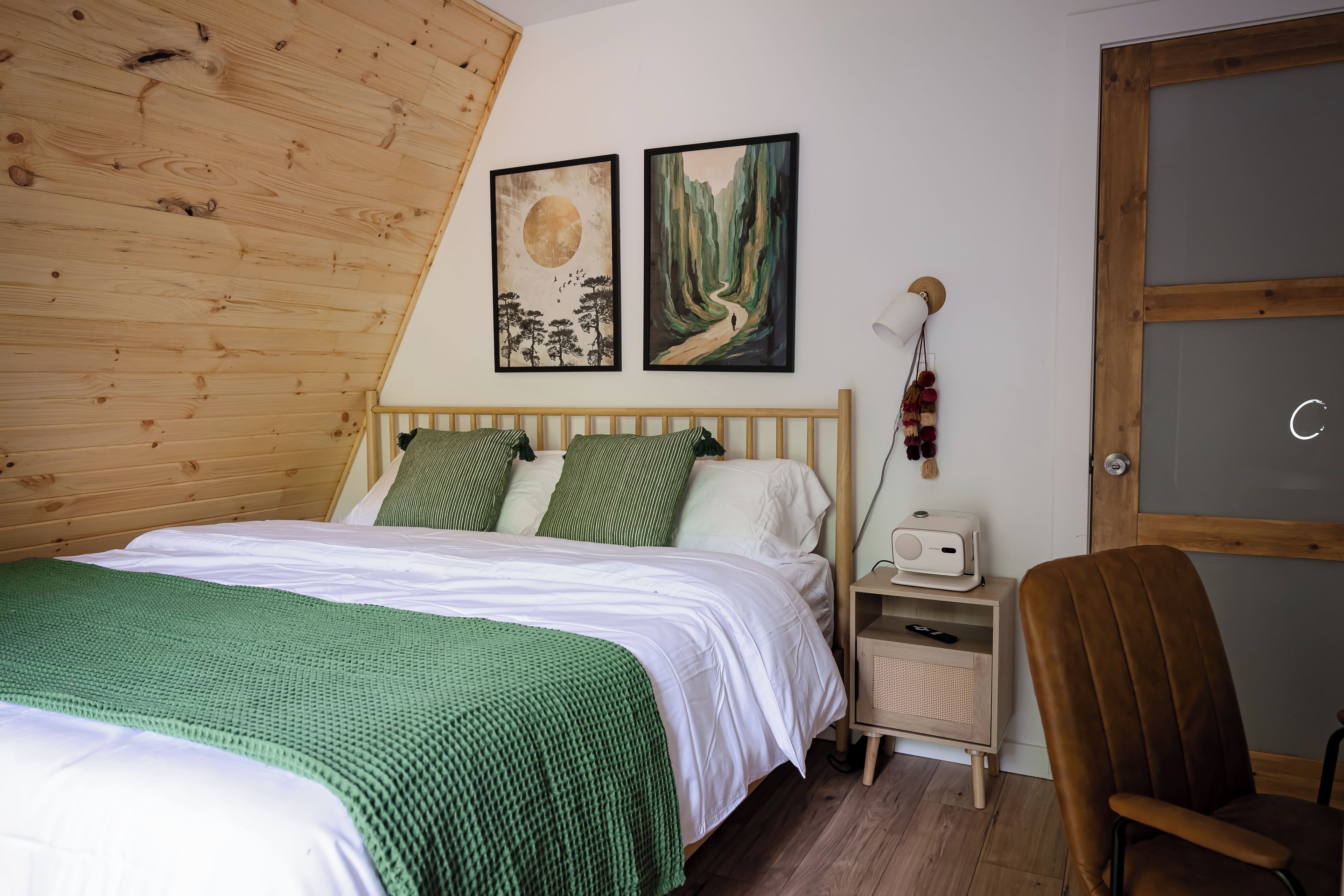 A bedroom with a wooden sloped ceiling, a bed with green and white bedding, two wall-mounted paintings, a bedside lamp, a chair, and a small bedside table with a lamp and a remote control.