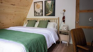 A bedroom with a wooden sloped ceiling, a bed with green and white bedding, two wall-mounted paintings, a bedside lamp, a chair, and a small bedside table with a lamp and a remote control. - Cozy A-frame w/HOT TUB, 5min walk to lake, King Bed, 30 min to Dells (Friendship)
