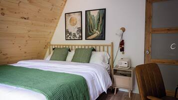 A bedroom with a wooden sloped ceiling, a bed with green and white bedding, two wall-mounted paintings, a bedside lamp, a chair, and a small bedside table with a lamp and a remote control.