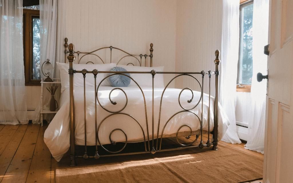 Quarto com cama de estrutura metálica, roupa de cama branca e janela com cortinas.