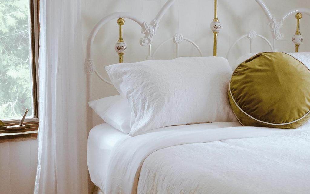 A bed with white bedding and a gold-colored pillow, a white metal headboard with ornate details, and a window with white curtains.