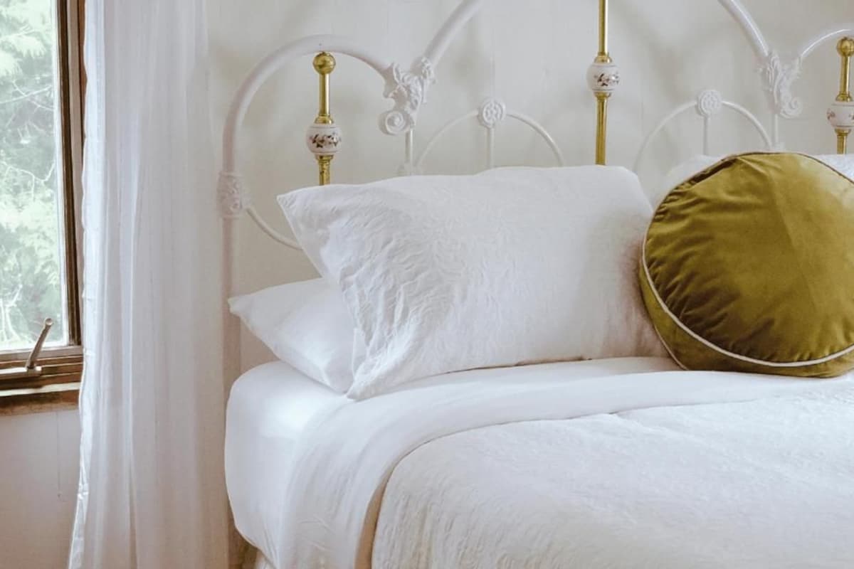 Un letto con biancheria bianca e un cuscino dorato, una testiera in metallo bianco con decorazioni elaborate, e una finestra con tende bianche.