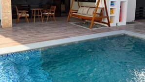 Une piscine avec une terrasse en bois, un fauteuil suspendu et un espace détente comprenant des chaises et une table.