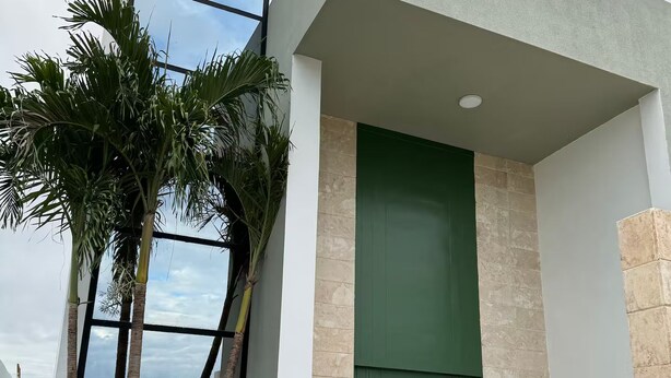 Un bâtiment moderne doté d’une grande fenêtre, d’une porte verte et d’un palmier devant l’entrée.