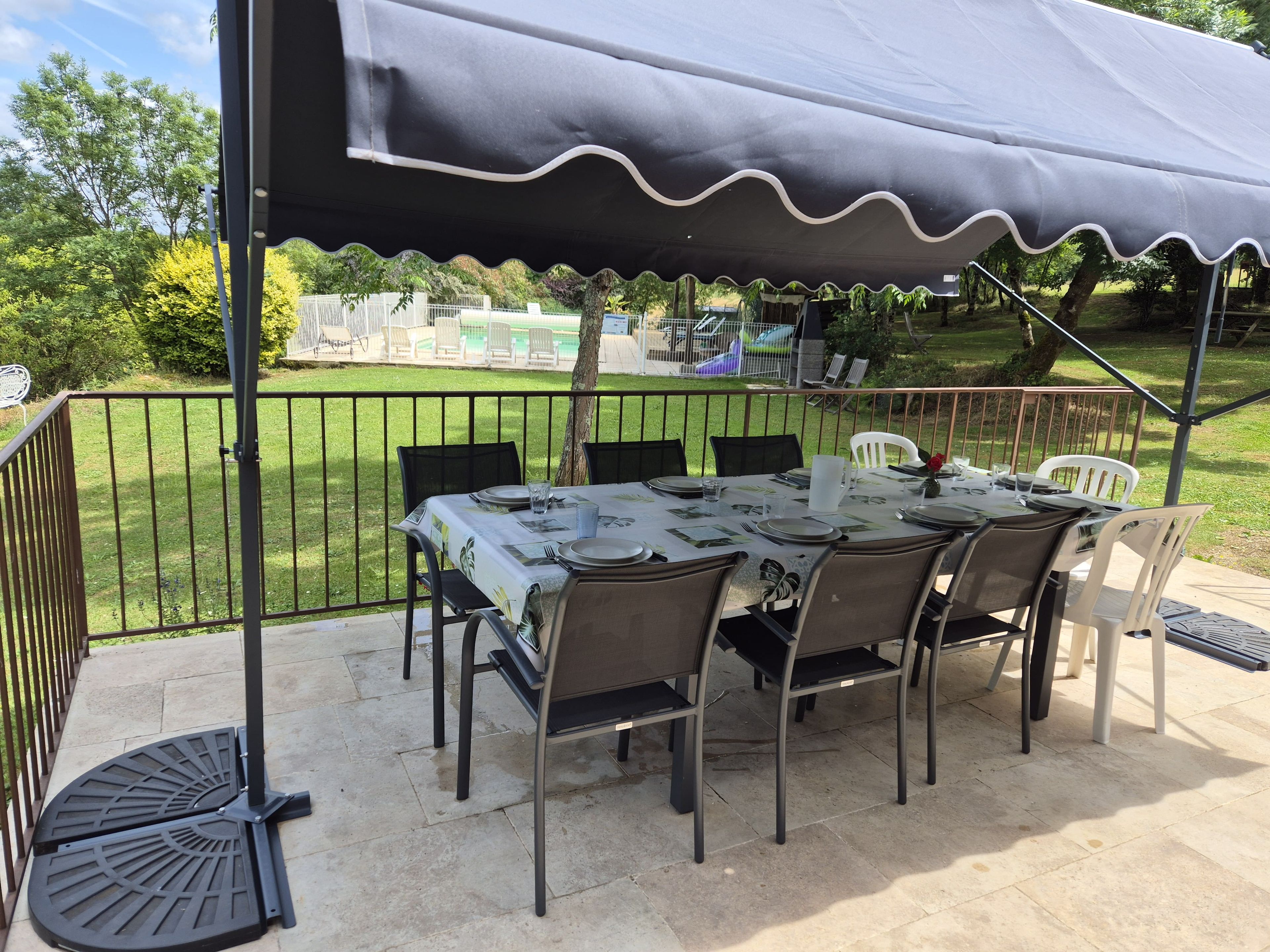 Un patio exterior con mesa lista para comer, rodeado de sillas, bajo una zona cubierta con vistas a una piscina y jardín.