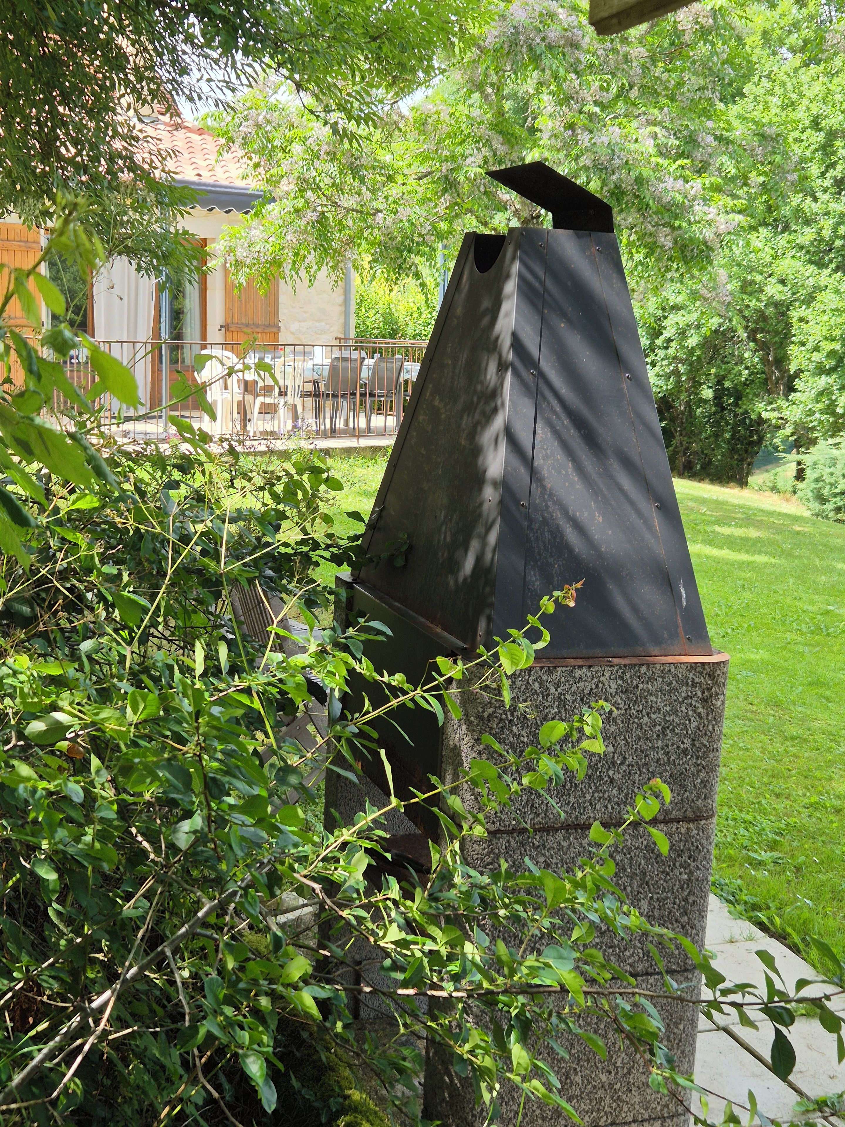 Un jardín con una estructura metálica, posiblemente una parrilla, rodeado de vegetación y un edificio con tejado de tejas al fondo.
