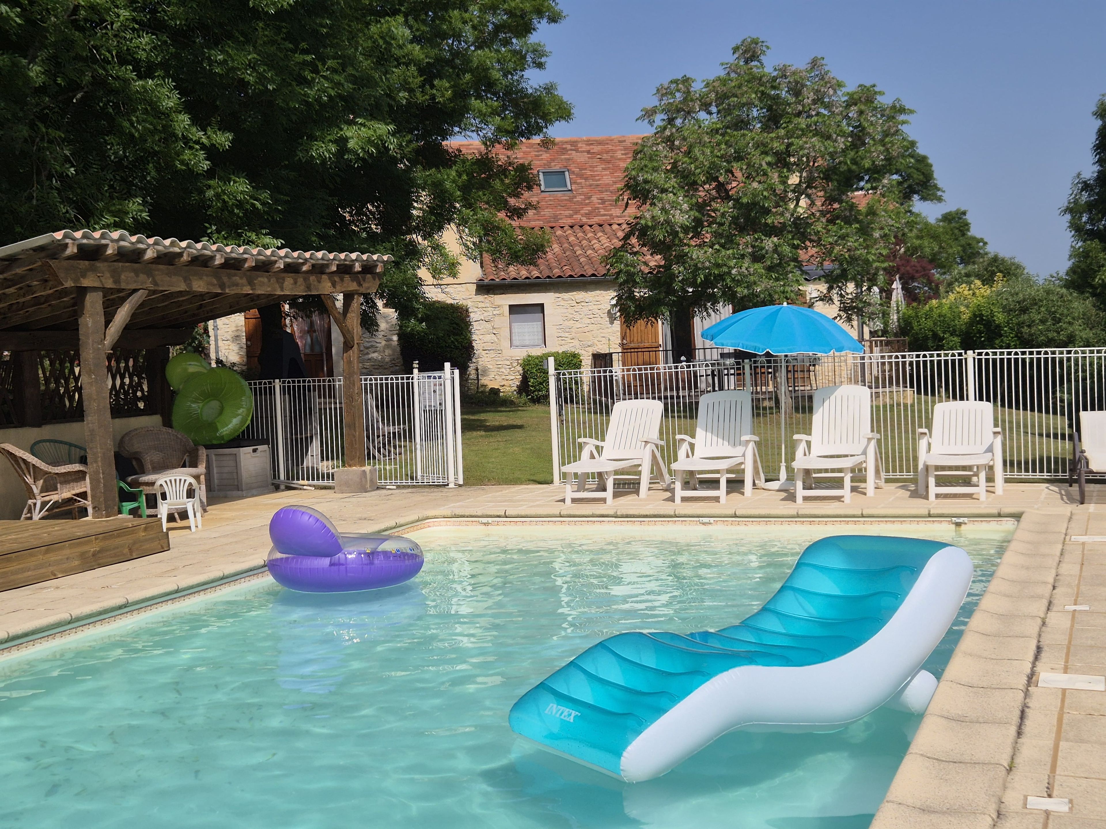 A swimming pool with inflatable loungers, a covered seating area, and a fenced backyard with lawn chairs.