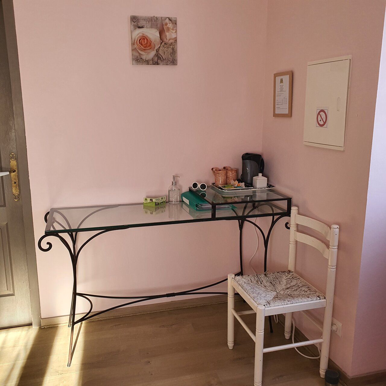 A small room with a glass-top table, a white chair, a framed picture on the wall, and a door with a lock.