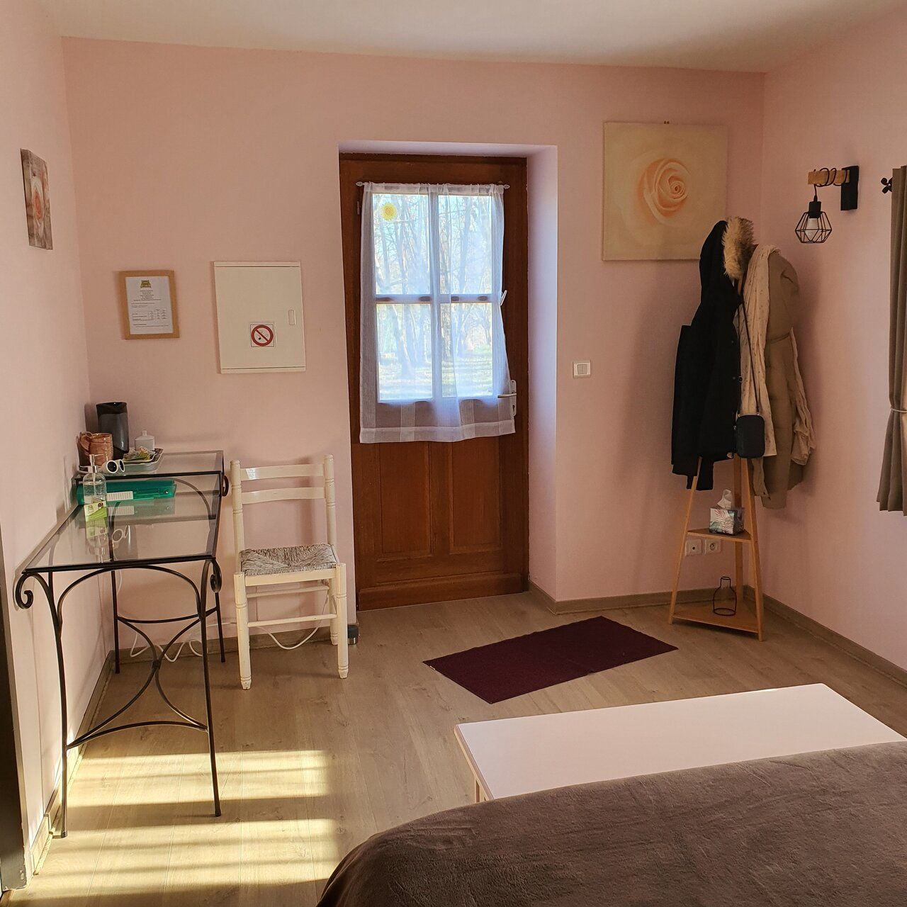 Una habitación con puerta de madera, una mesa con sillas, un perchero con ropa y una cama.