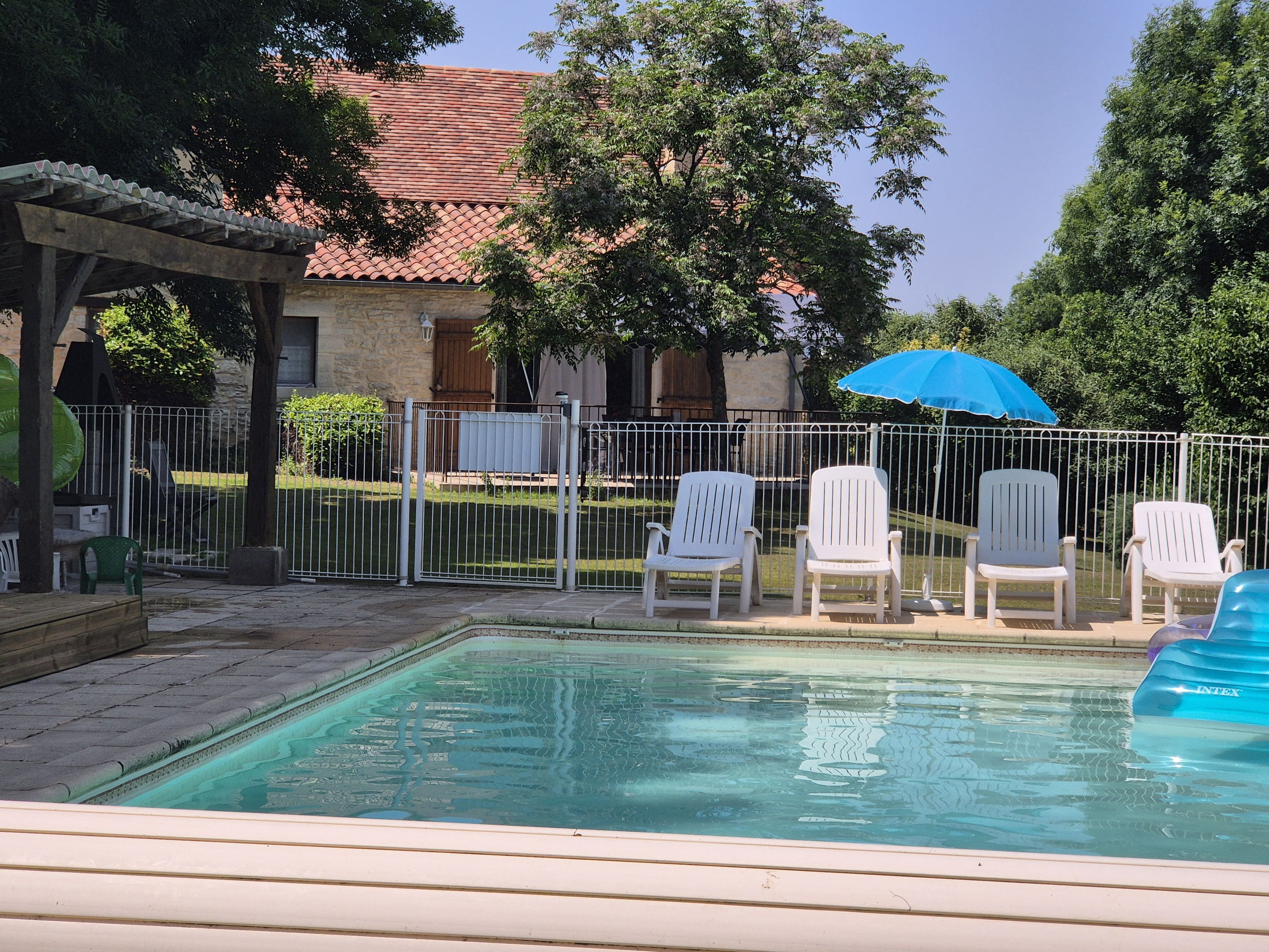 Une piscine entourée d’une clôture et d’arbres, avec des chaises blanches et un parasol bleu.