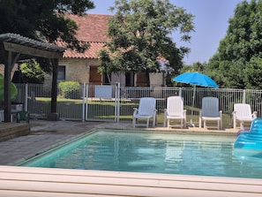 A swimming pool with white chairs and a blue umbrella, surrounded by a fence and trees.