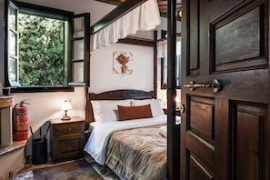 Een slaapkamer met een groot bed, een nachtkastje met een lamp, een raam met uitzicht op bomen, en een deur met een deurklink.