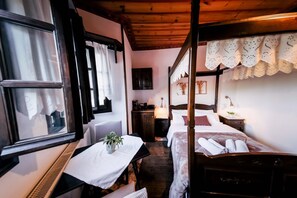 Ein traditionell eingerichteter Raum mit einem Himmelbett, einem kleinen Tisch mit einer Pflanze und einem Fenster mit Vorhängen.