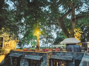 An outdoor seating area with tables and chairs under large trees, a covered area, and stone structures.