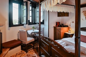 A bedroom with a four-poster bed, a desk with a chair, a bedside table with a lamp, and a view through the window.
