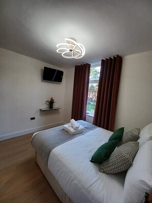 A hotel room with a bed, a flat-screen TV mounted on the wall, a window with curtains, and a small plant on a shelf. - International House (London)