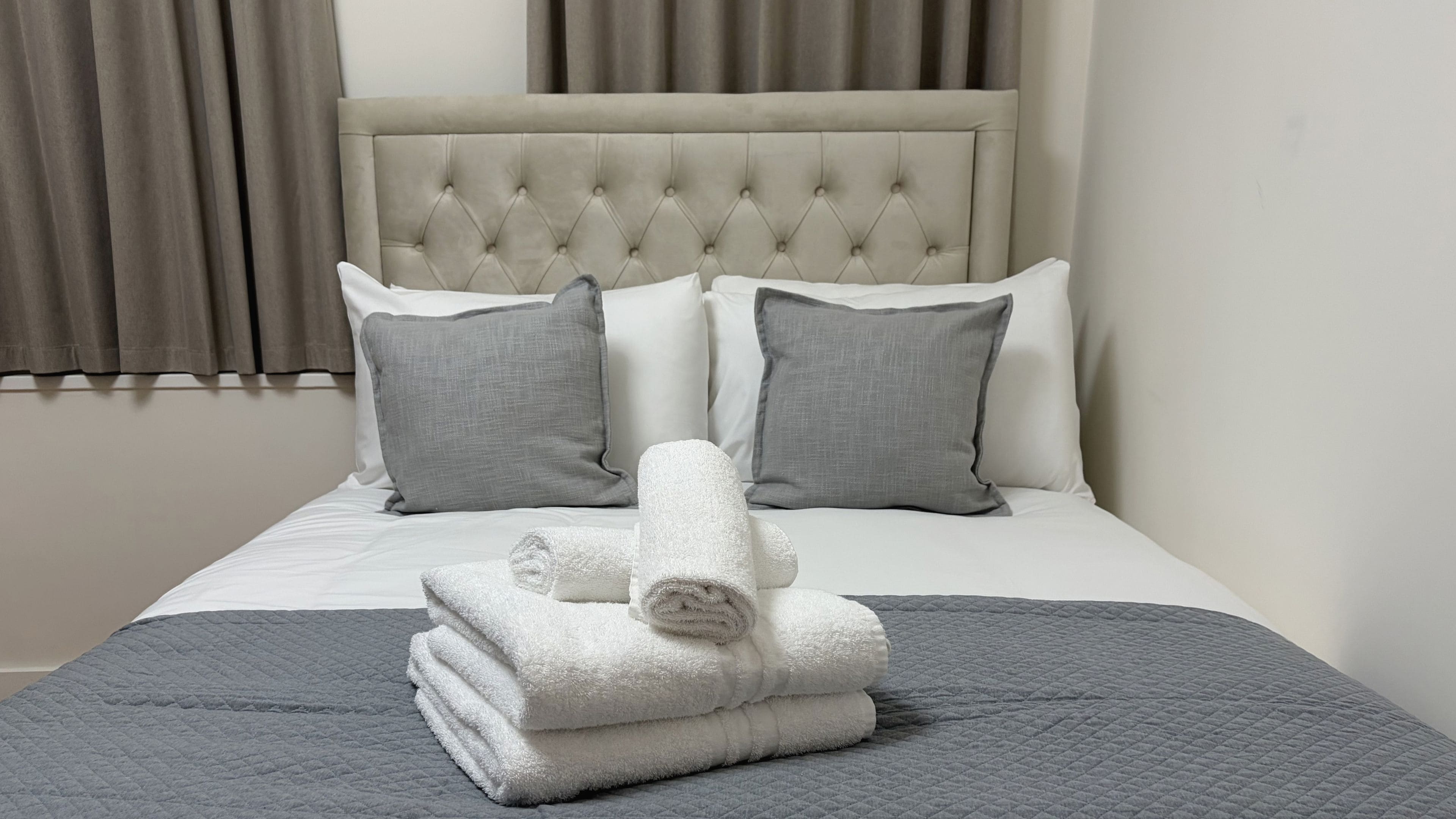 Una cama bien hecha con colcha gris, dos almohadas grises y una toalla blanca doblada.