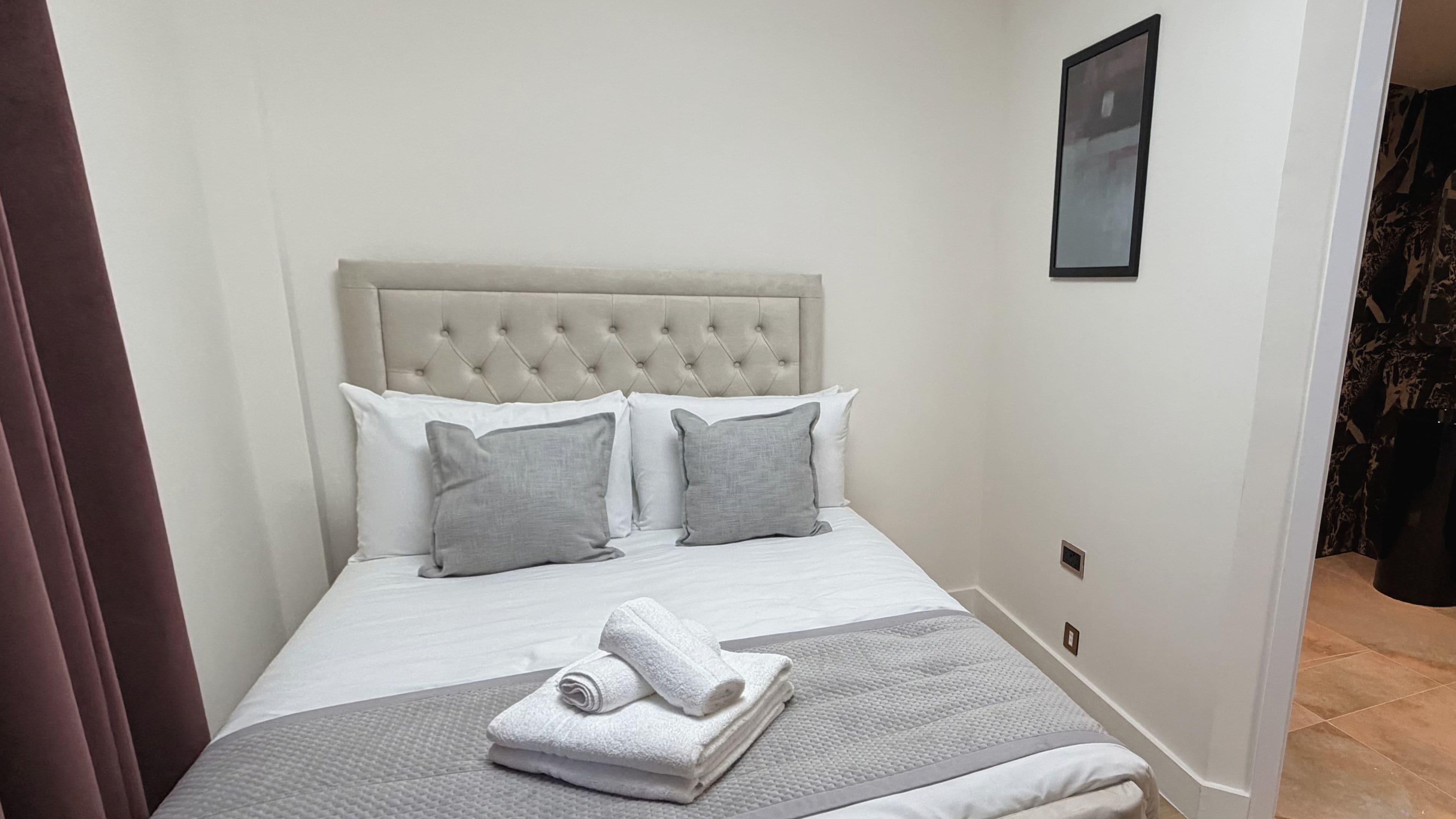 A neatly made bed with a tufted headboard, white and grey pillows, and a folded towel on a grey textured blanket.