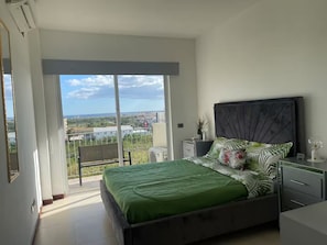 Un dormitorio con cama, mesita de noche, cómoda, planta en maceta y vista a la ciudad a través de amplias ventanas.