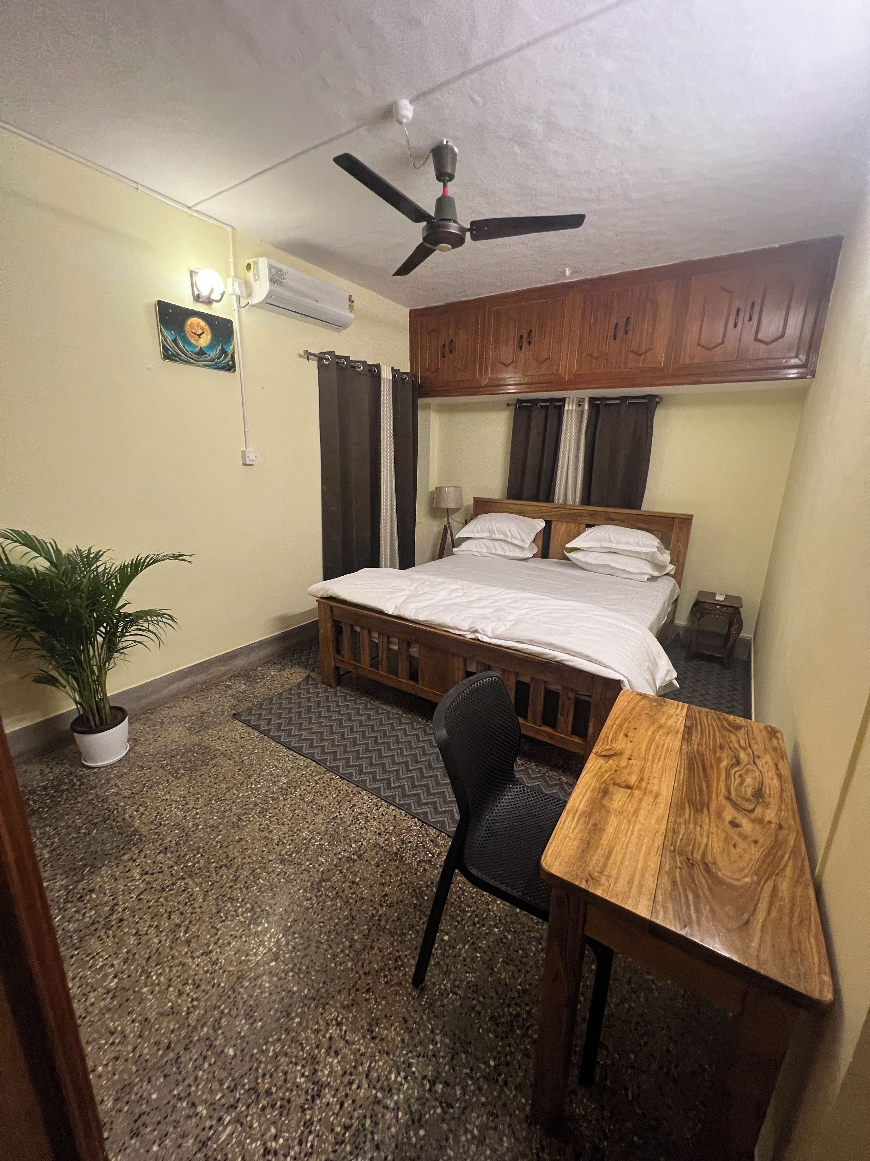 A bedroom with a bed, a desk, a chair, a ceiling fan, a potted plant, and a wall-mounted air conditioner.
