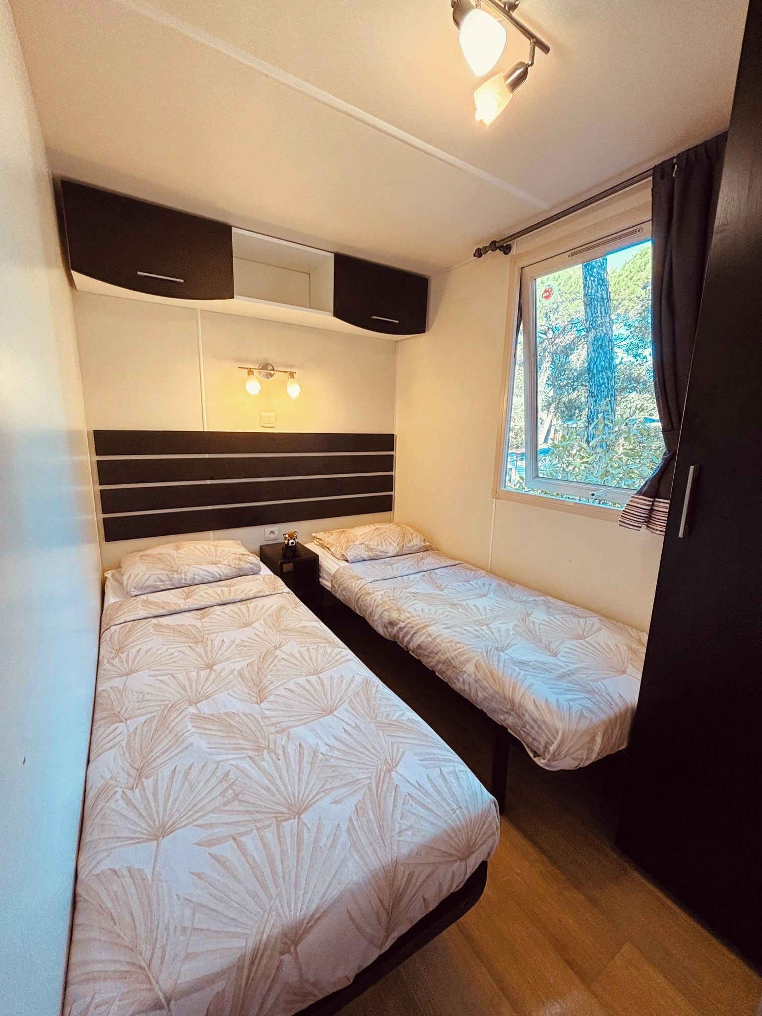 A compact room with two beds, a window with curtains, and a ceiling light fixture.