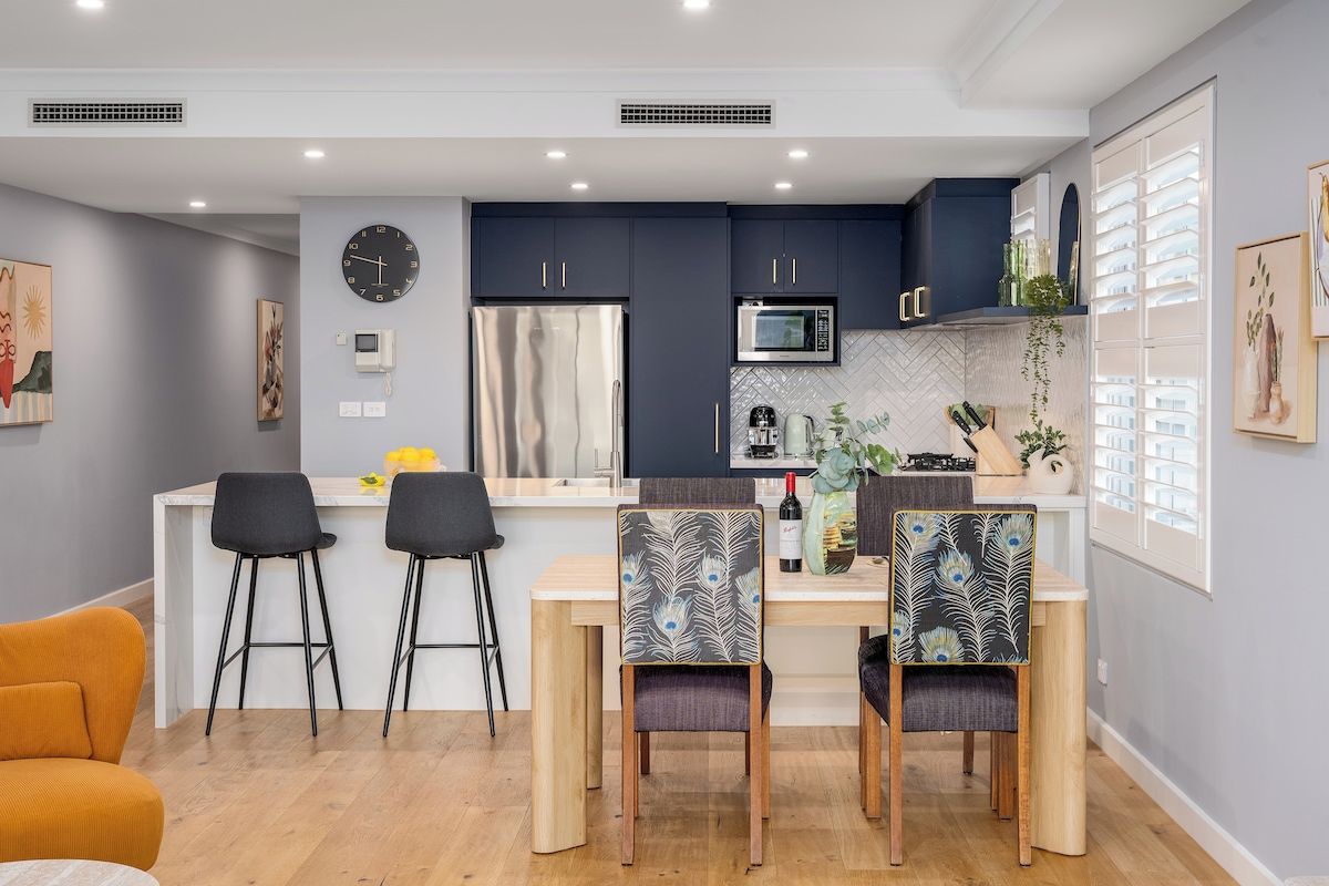 A modern kitchen with dark cabinets, a microwave, and a refrigerator. There are two bar stools and a dining table with chairs.