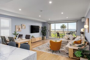 A modern living room with a dining area, a sofa, and a TV mounted on the wall. - Beachfront 3BR Retreat w Parking, Pool and Tennis (Saint Kilda West)