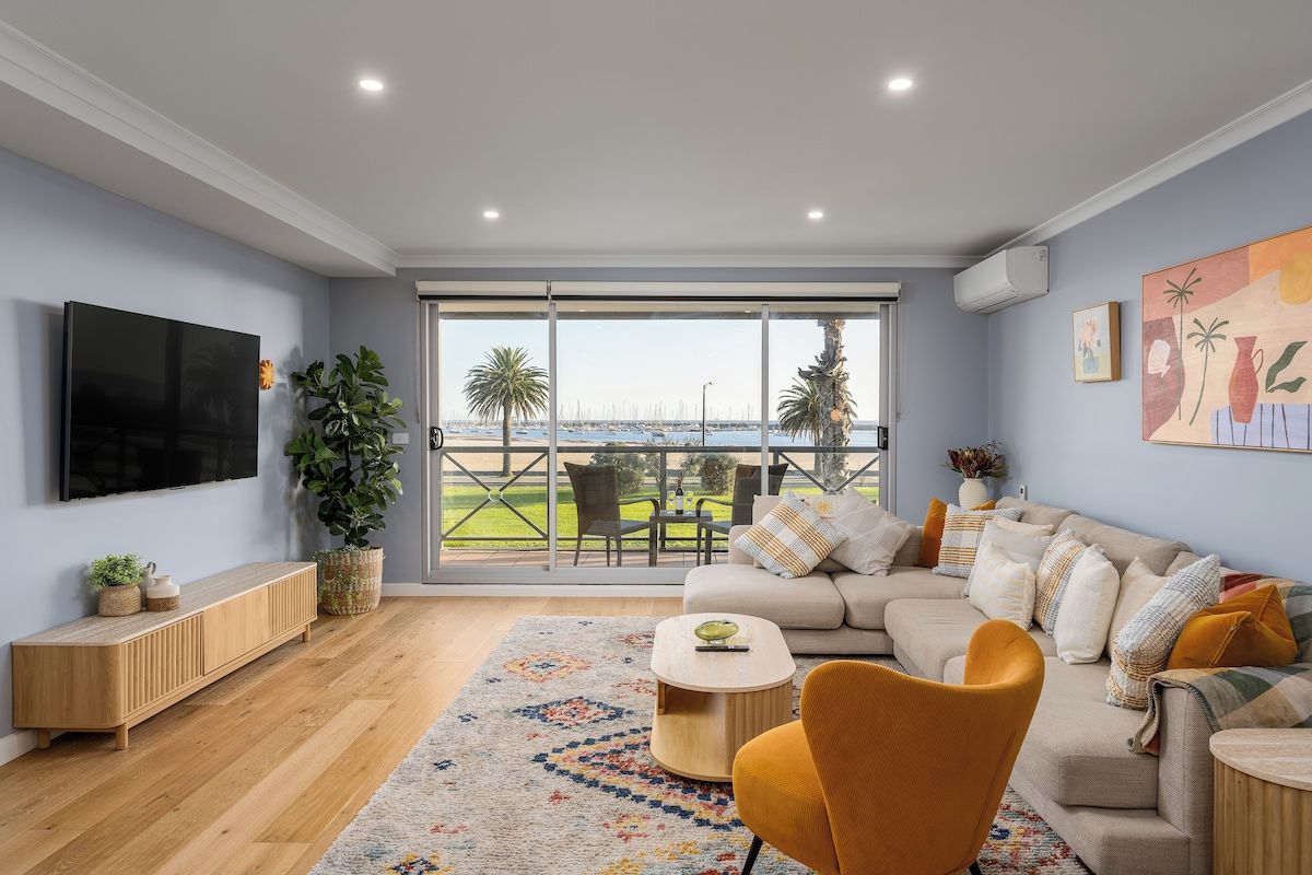 A modern living room with a large TV, a sofa, a coffee table, and a view of a marina through large windows.