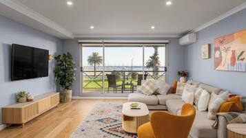 A modern living room with a large TV, a sofa, a coffee table, and a view of a marina through large windows.