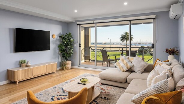 Una sala de estar moderna con un televisor grande, un sofá, una mesa de centro y vistas a un campo deportivo a través de ventanas amplias.