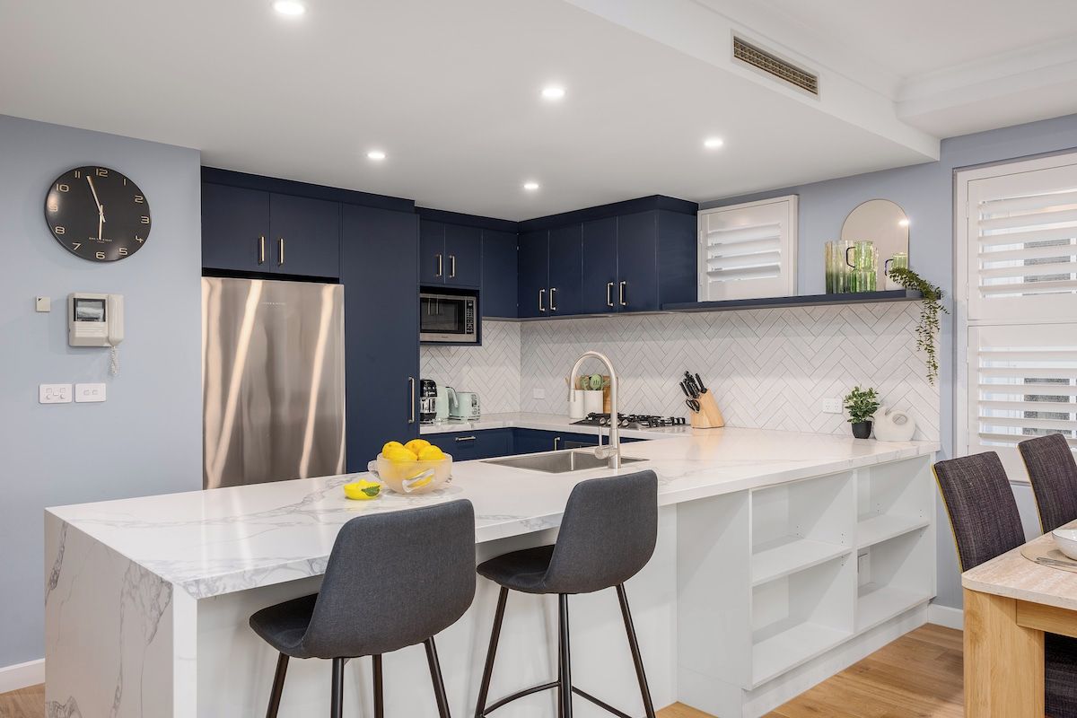A modern kitchen with dark blue cabinets, a stainless steel refrigerator, a microwave, and a sink. There is a marble countertop with two bar stools and a small plant on the counter.
