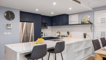 A modern kitchen with dark blue cabinets, a stainless steel refrigerator, a microwave, and a sink. There is a marble countertop with two bar stools and a small plant on the counter.