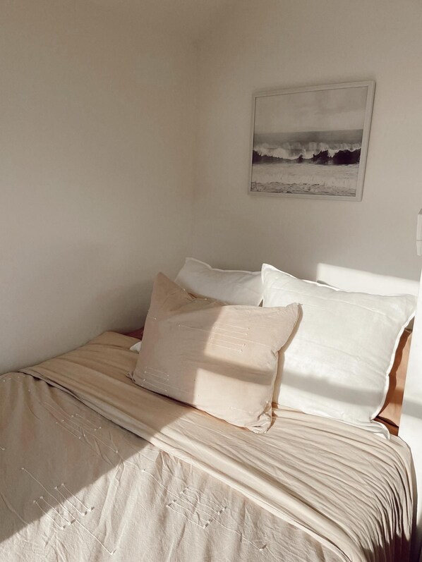 A bed with a beige bedspread and pillows, a framed picture of a beach scene on the wall, and a window allowing natural light into the room.