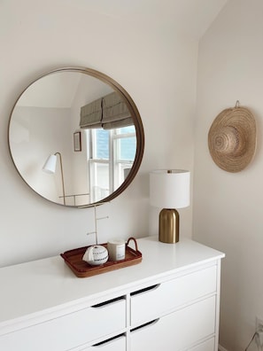 A white dresser with a round mirror, a lamp, and a woven basket with candles.