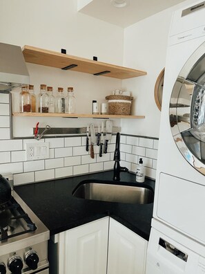 A kitchen with white cabinets, a black countertop, a stainless steel sink, and a stove. There are jars on a shelf above the sink and a basket on the countertop. - Cozy 2-bedroom Beach Bungalow in lovely Ocean Grove, NJ with AC (Neptune Township)