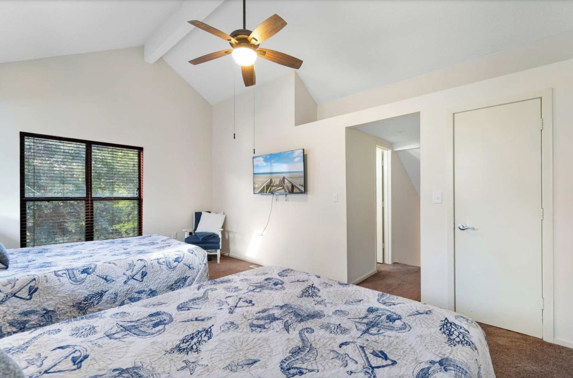A bedroom with two beds, a ceiling fan, a TV, and a window with blinds.