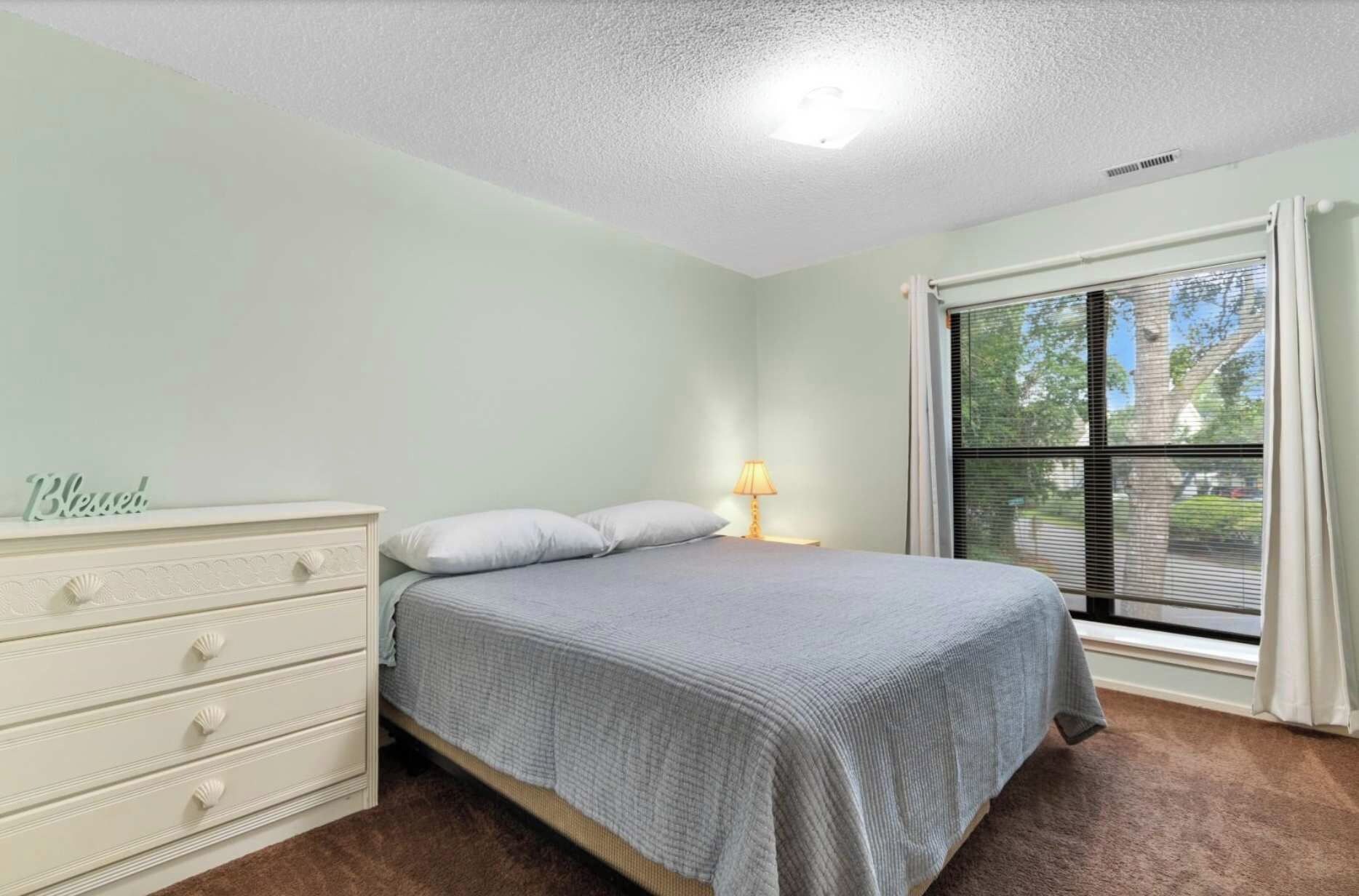 A bedroom with a bed, a dresser, a nightstand with a lamp, a window with blinds, and a ceiling light.
