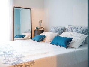 Ein ordentlich bezogenes Bett in Weiß und Blau, ein Nachttisch mit Lampe und ein Spiegel an der Wand.