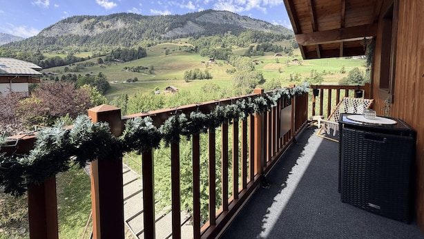 Un balcone con vista su una valle verde e montagne, ringhiere in legno e una sedia in vimini.