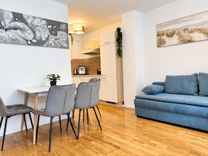 Una sala de estar moderna con un sofá azul, sillas grises, piso de madera y una cocina al fondo.