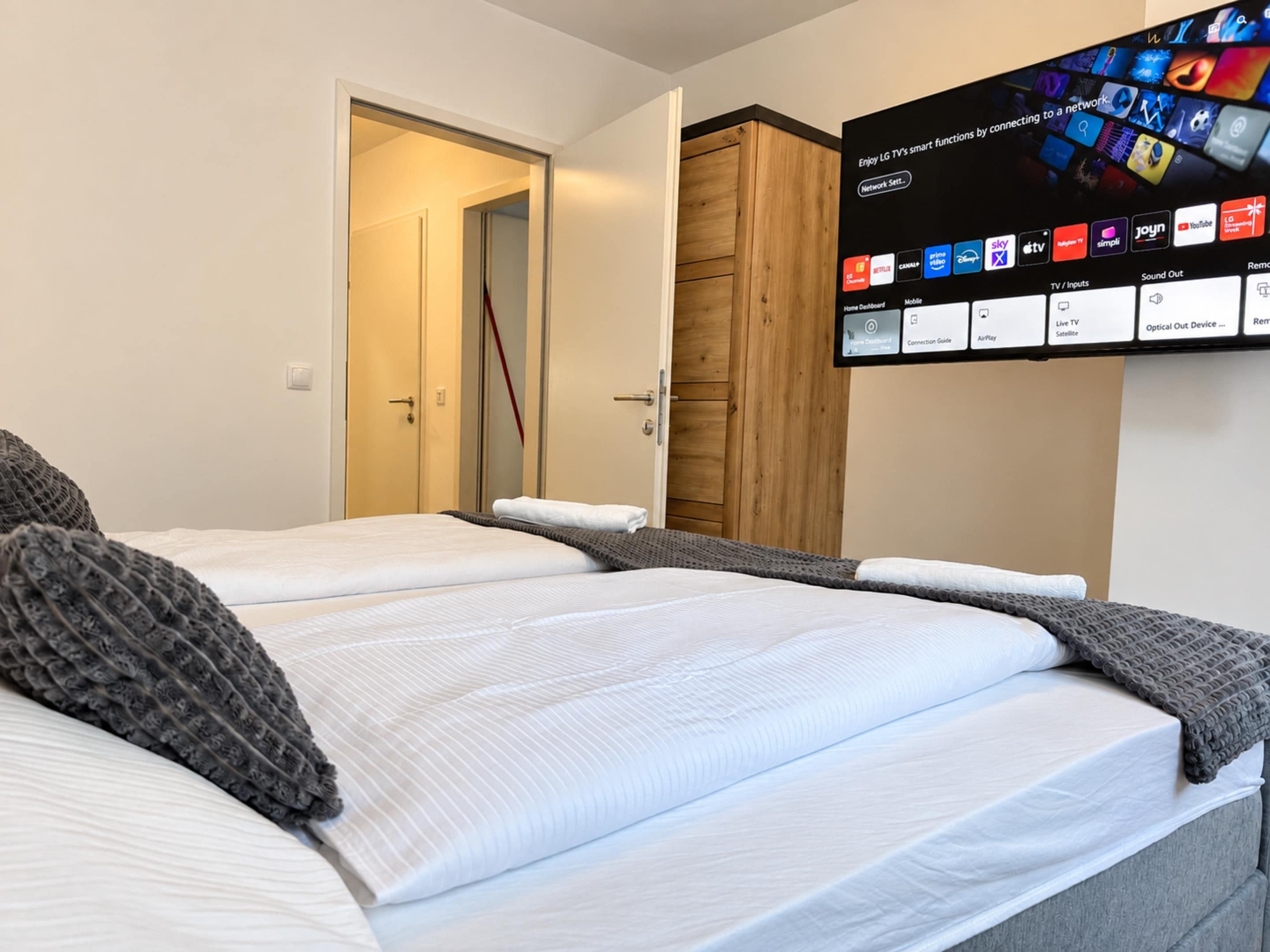 A hotel room with two beds, a TV mounted on the wall displaying channel options, and a wooden wardrobe.