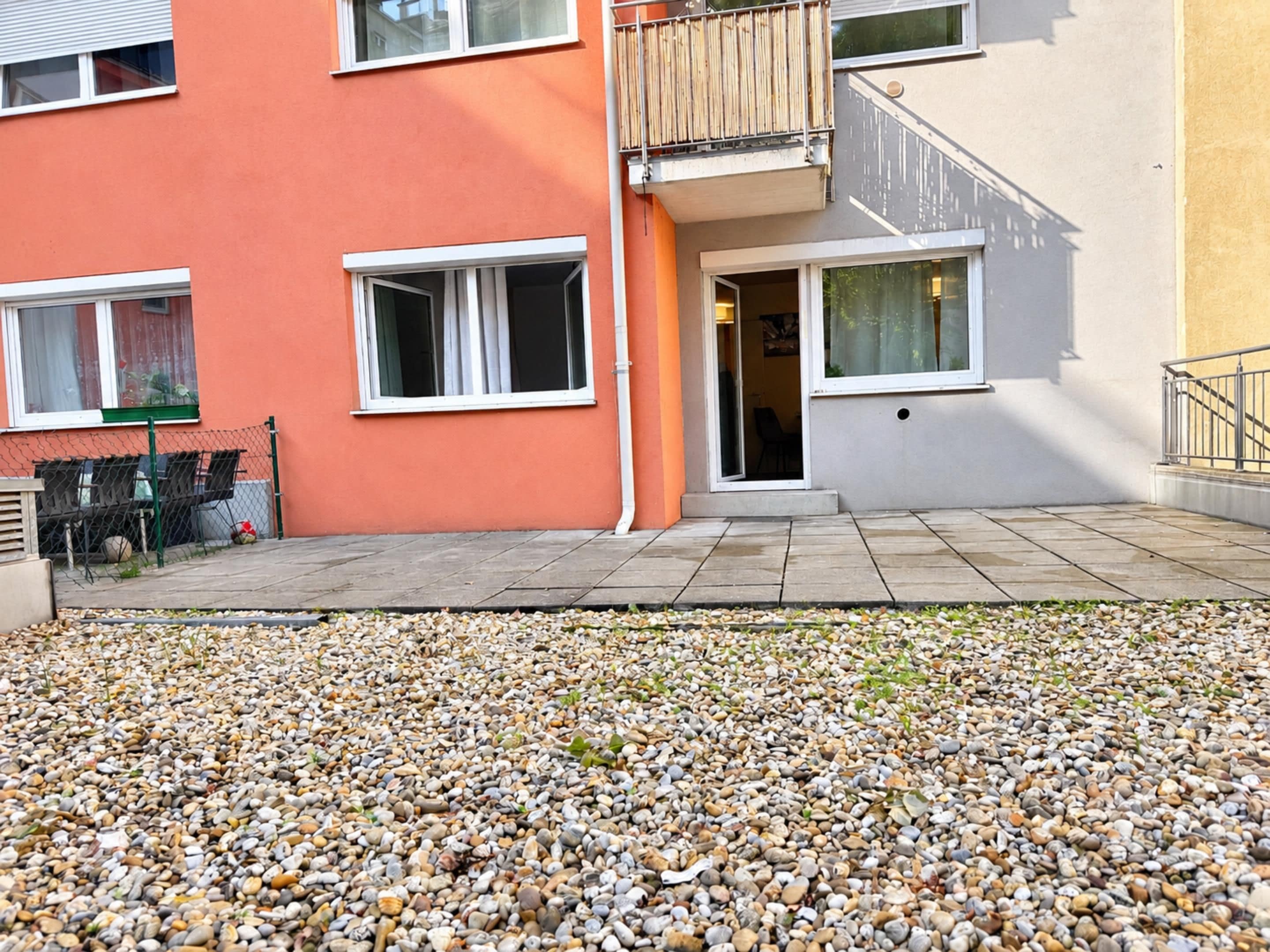 A residential building with a pebble-covered ground, a balcony, and a staircase leading to an entrance.