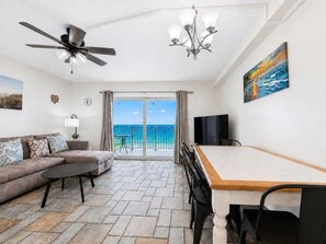 Ein Zimmer mit einer Couch, einem Esstisch, einem Fernseher, einem Deckenventilator, einer Kandelaber und einem Gemälde mit Strandmotiv.