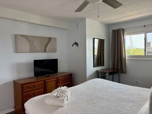 Ein Hotelzimmer mit einem Bett, einem Fernseher auf einer Holzkommode, einem Deckenventilator, einem Fenster mit Vorhängen und einem Wandbild.
