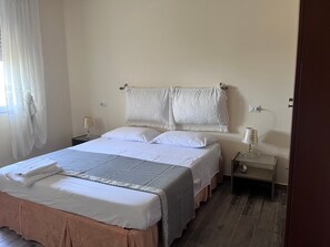 A neatly made bed with white linens and a pink skirt, two bedside tables with lamps, and a window with sheer curtains.