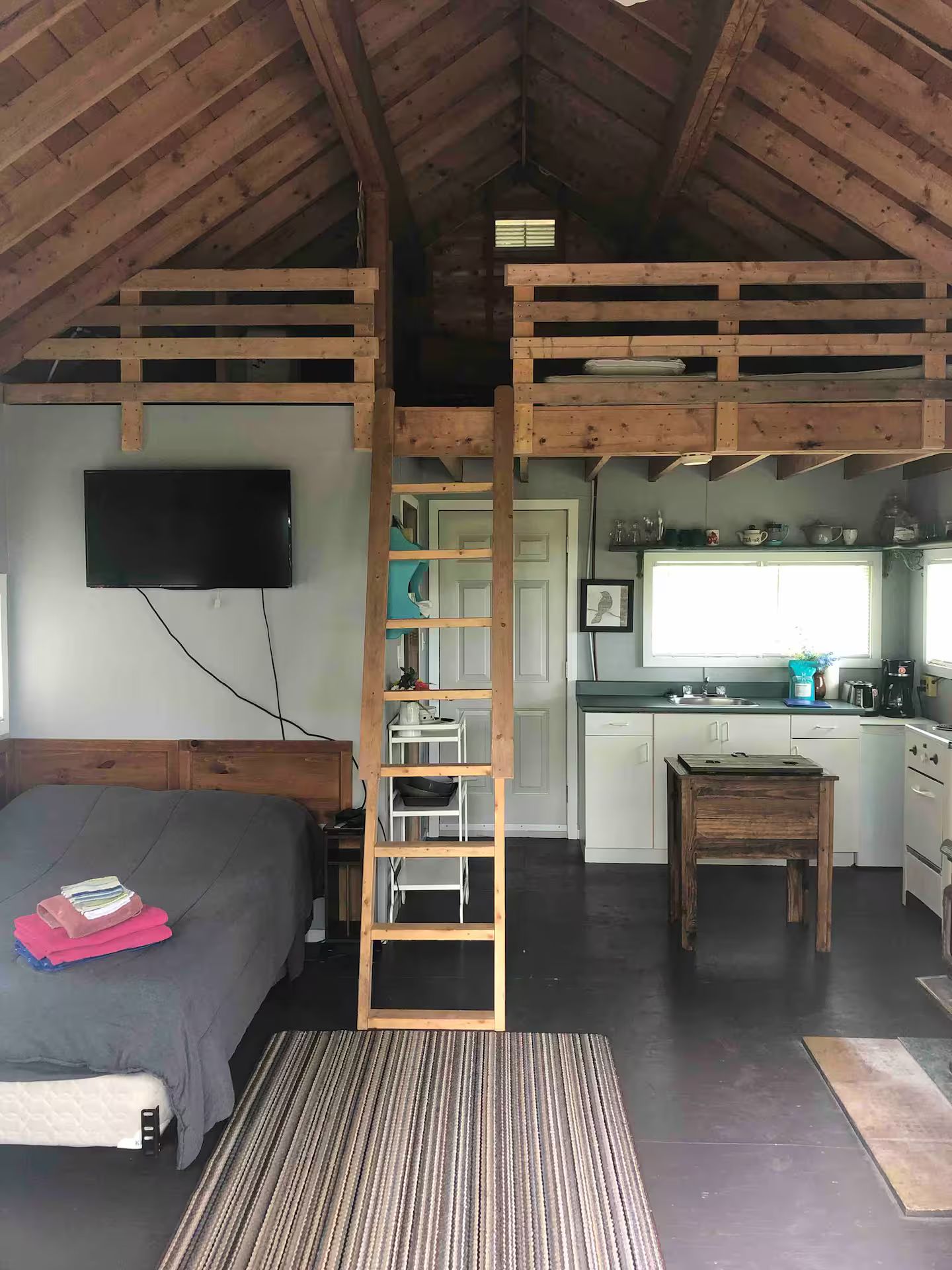 Habitación estilo loft con cama, una escalera que lleva a una zona elevada, cocina y televisor montado en la pared.